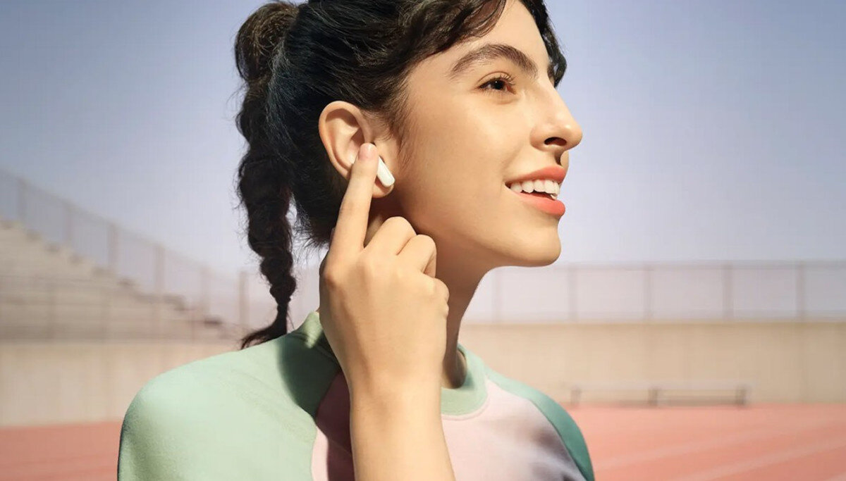 Osoba nosi białe słuchawki dokanałowe na tle stadionu. Ubrana jest w kolorową bluzę z krótkim rękawem.