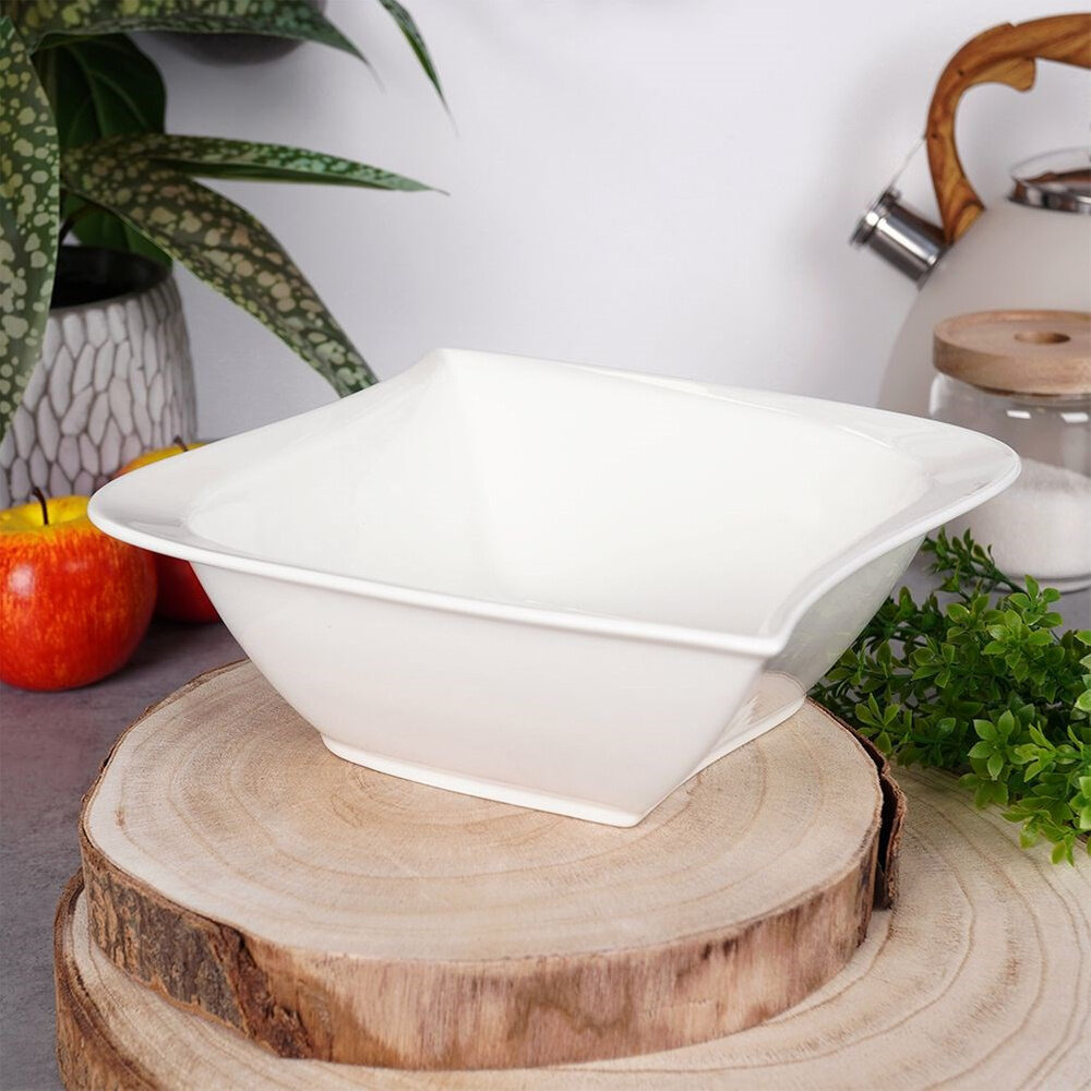 Biała porcelanowa miska sałatkowa o kwadratowym kształcie, umieszczona na drewnianej podstawce. W tle widoczne są rośliny, jabłka i czajnik.