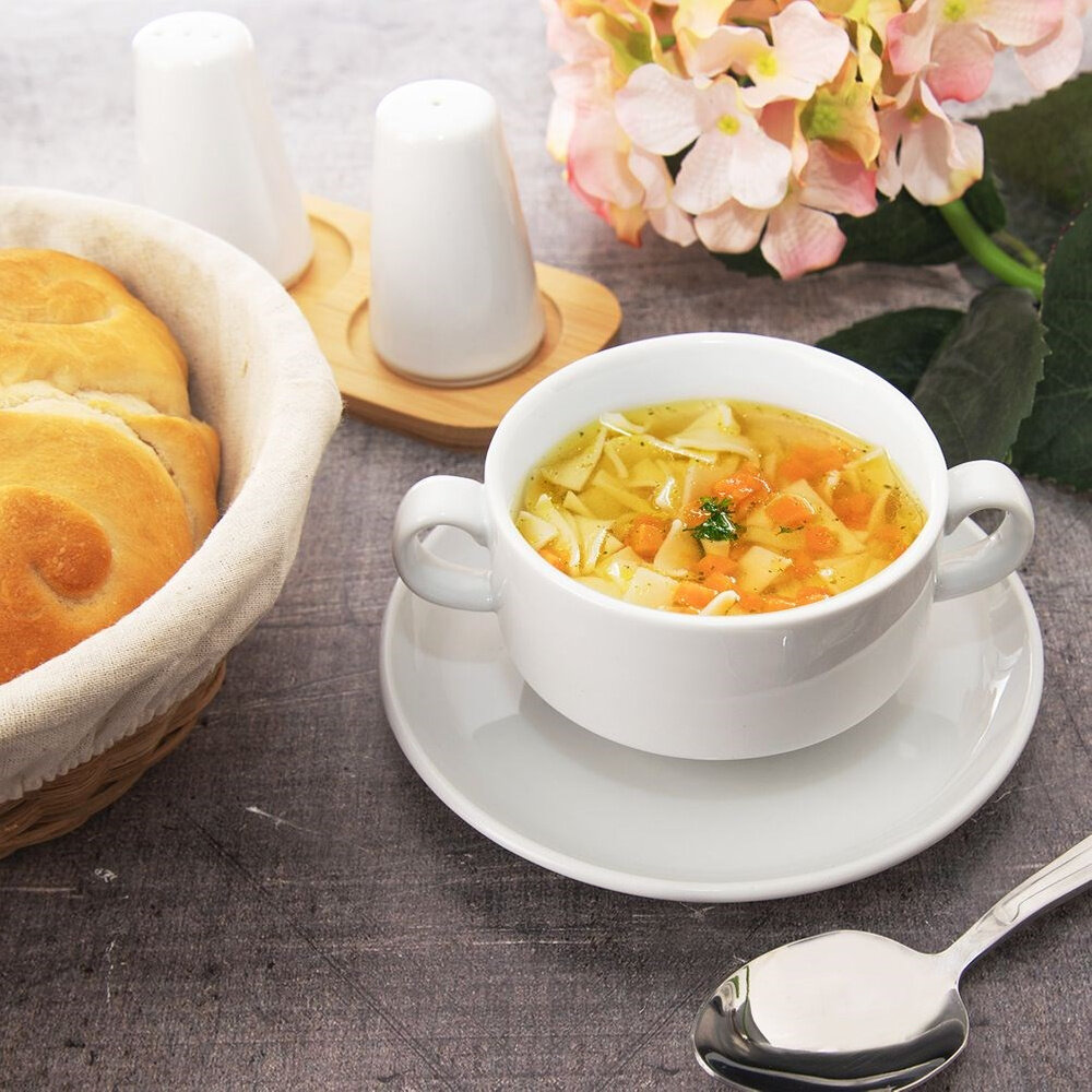 Na obrazie widoczna jest porcelanowa miska z bulionem, obok której leży łyżka. W tle znajduje się koszyk z pieczywem, solniczka i pieprzniczka oraz kwiaty.