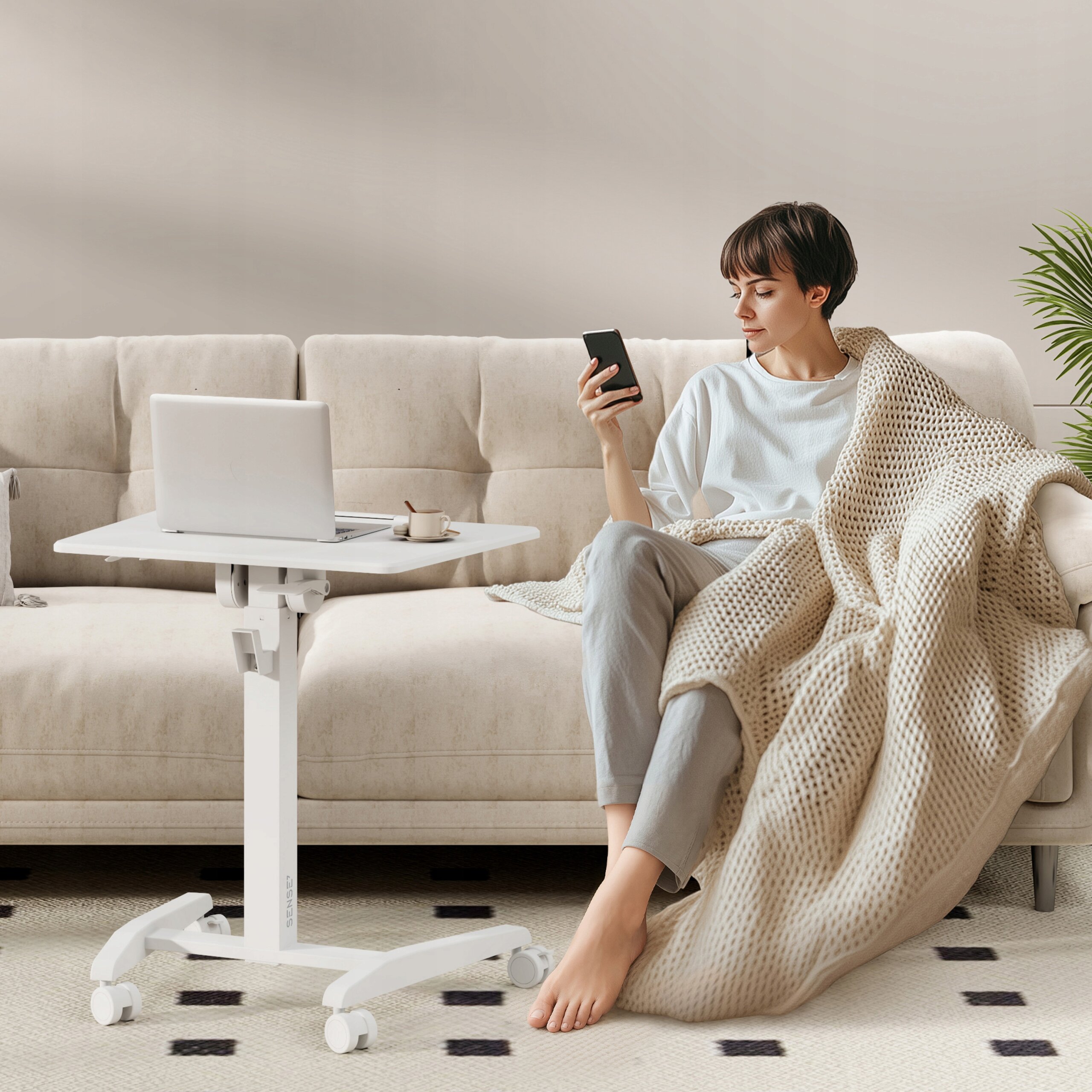 Osoba siedzi na kanapie, trzymając telefon, obok znajduje się białe biurko mobilne z laptopem i filiżanką. W tle widać jasną ścianę i roślinę doniczkową.