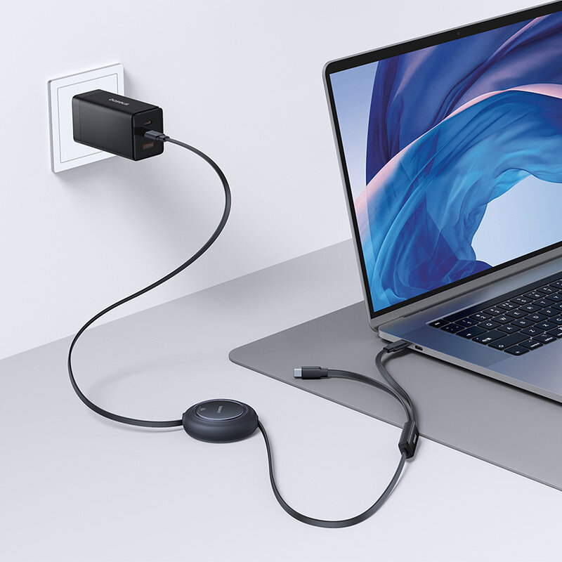Na obrazie widać laptop podłączony do gniazdka elektrycznego za pomocą czarnego kabla USB-C, który jest zwijany w okrągłym mechanizmie. Na ładowarce widoczne jest logo \'Baseus\'.