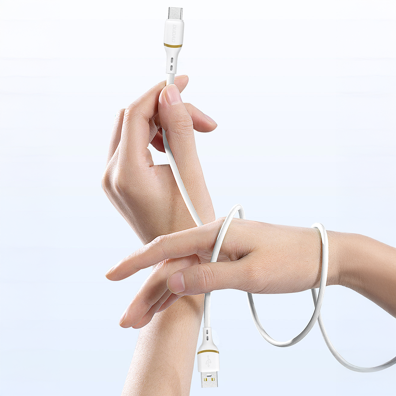 Na zdjęciu widać białe ręce trzymające biały kabel USB z końcówkami USB-A i Lightning. Kabel jest owinięty wokół nadgarstka, a na końcówce widoczne jest logo producenta Dudao.