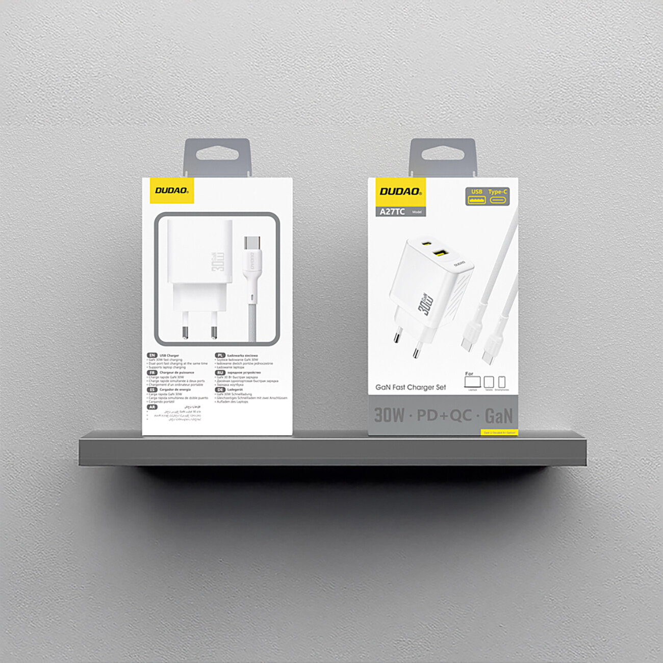 Na obrazie widoczne są dwa opakowania produktu marki Dudao, przedstawiające ładowarkę sieciową z kablem USB-C. Opakowania mają biało-szary design z żółtymi akcentami i zawierają teksty: \'USB\', \'Type-C\', \'30W\', \'PD+QC\', \'GaN\'.