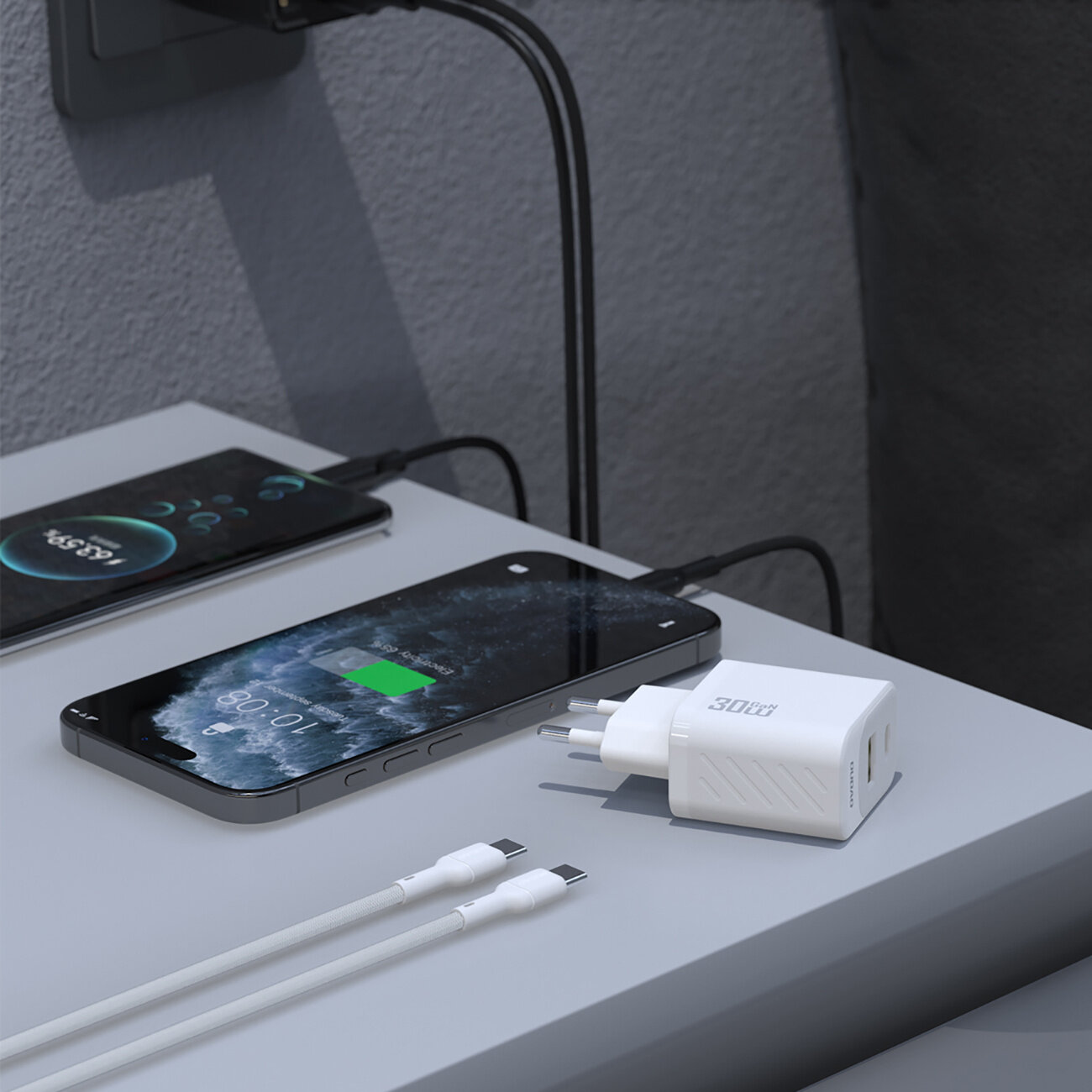 Na zdjęciu widać białą ładowarkę sieciową z dwoma portami USB-A i USB-C oraz kablem USB-C. Obok ładowarki leży smartfon z widocznym ekranem ładowania, a w tle widać drugi telefon podłączony do gniazdka.