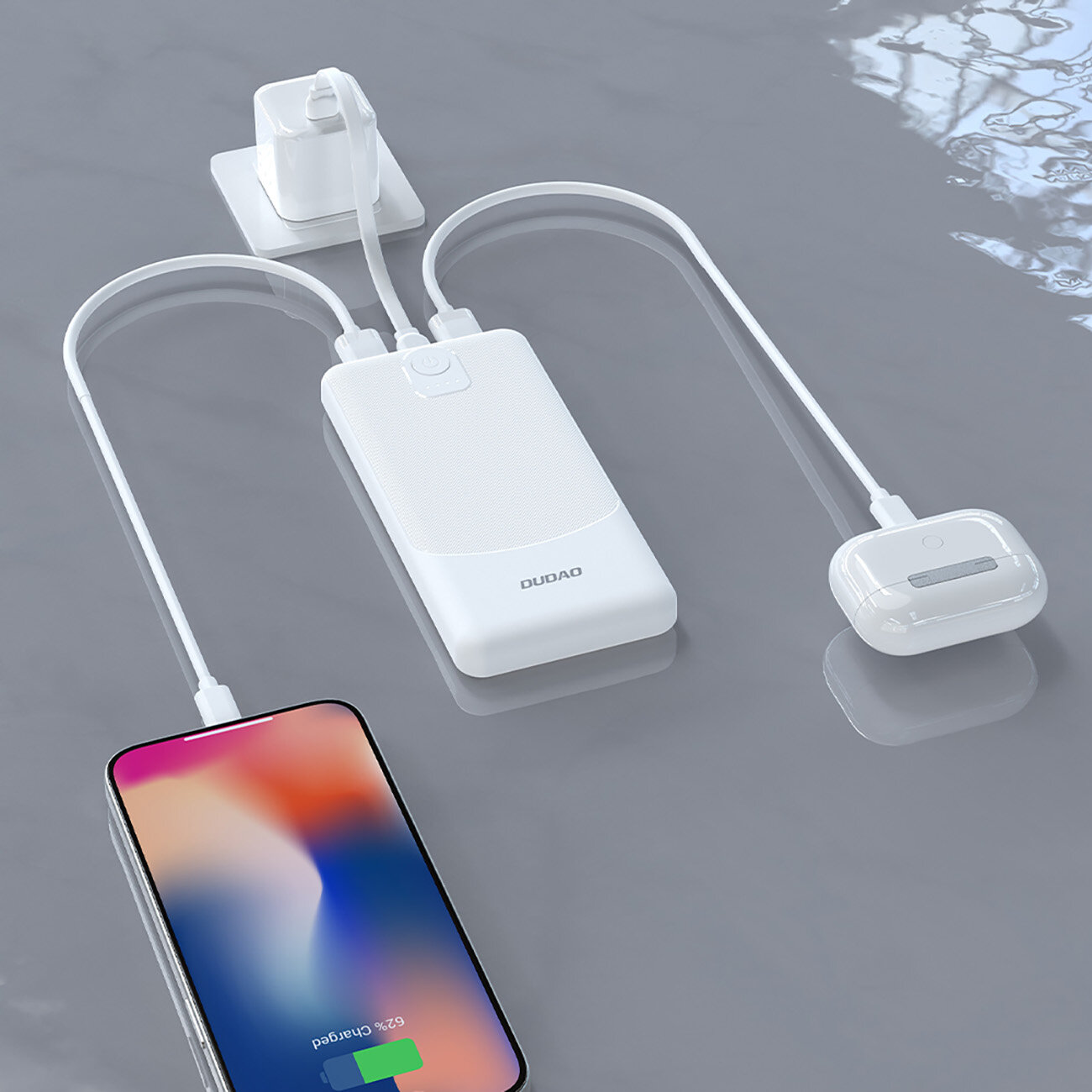 Na obrazie widoczny jest biały powerbank z napisem \'DUDAO\', do którego podłączone są dwa urządzenia za pomocą kabli USB. Obok znajduje się smartfon z wyświetlaczem pokazującym 62% naładowania baterii.
