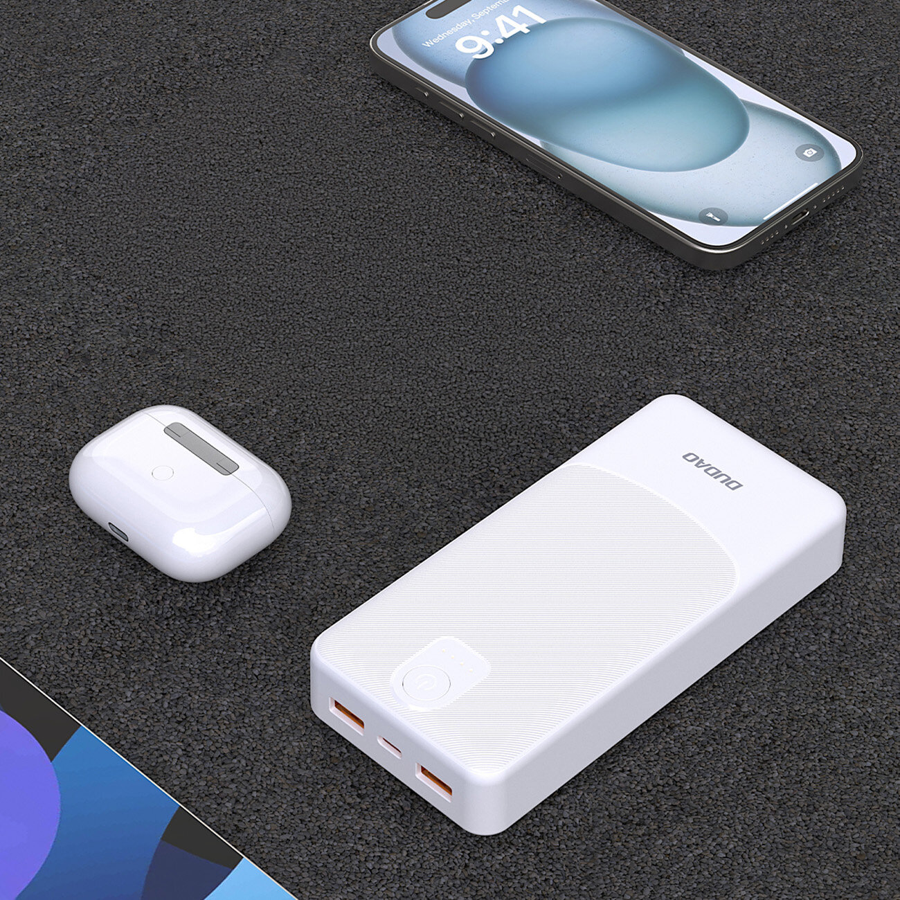 Na ciemnym tle znajduje się biały powerbank z dwoma portami USB-A i napisem \'DUDAO\'. Obok leży smartfon z wyświetlaczem oraz białe etui na słuchawki bezprzewodowe.
