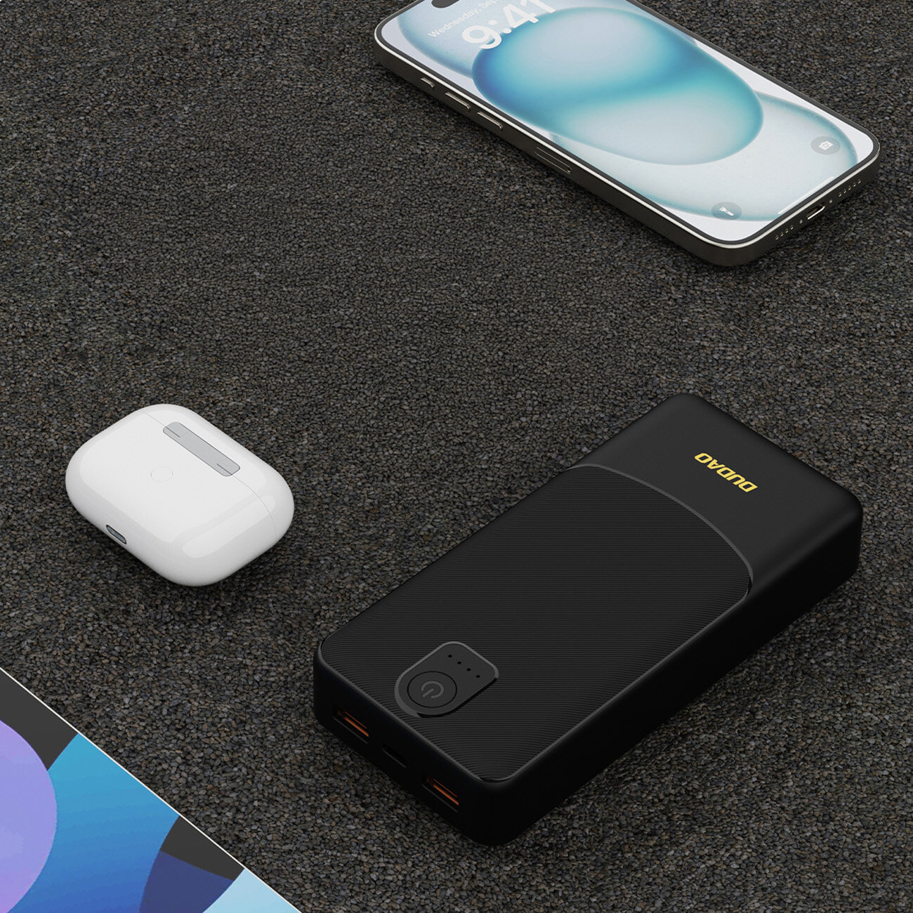 Na ciemnym tle leży czarny powerbank z dwoma portami USB-A i napisem \'DUDAO\'. Obok znajdują się białe słuchawki bezprzewodowe i smartfon z niebieskim ekranem.