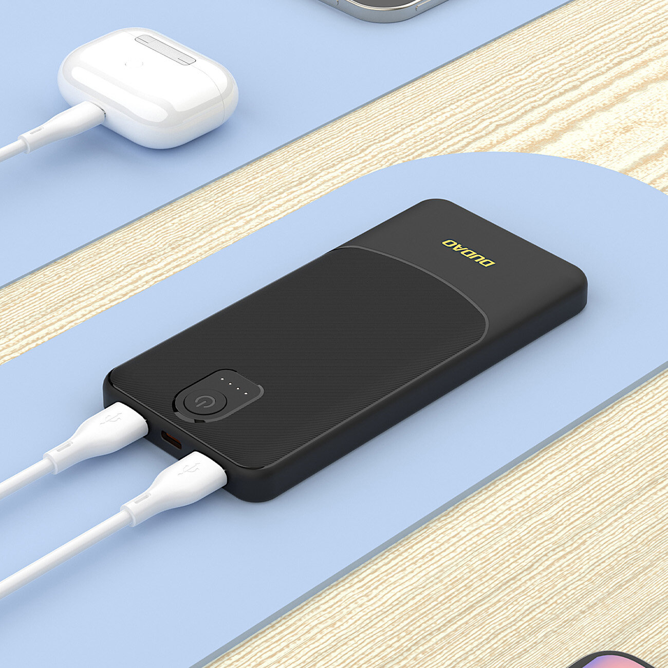Czarny powerbank z logo \'DUDAO\' na górze, z dwoma podłączonymi białymi kablami USB. W tle widoczne są inne urządzenia elektroniczne na jasnym drewnianym blacie.