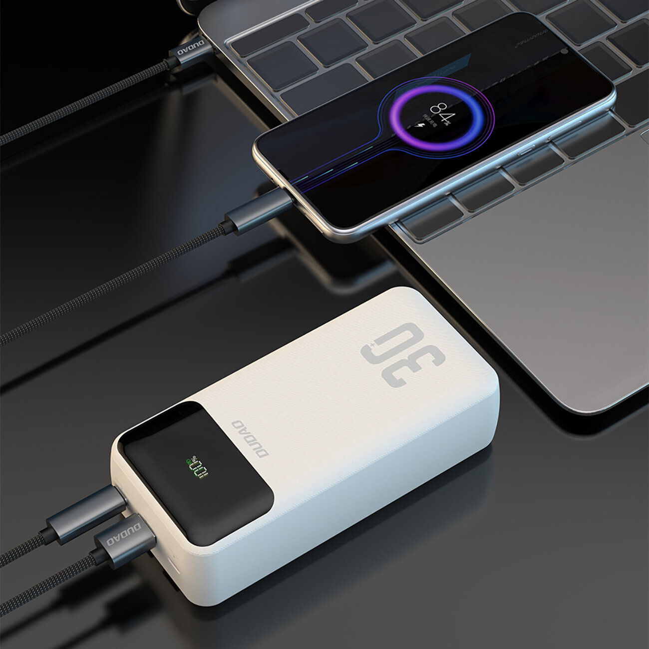 Na obrazie widoczny jest biały powerbank z wyświetlaczem pokazującym \'100%\' oraz napisem \'DUDAO\'. Obok znajduje się smartfon podłączony do laptopa za pomocą kabla USB-C.