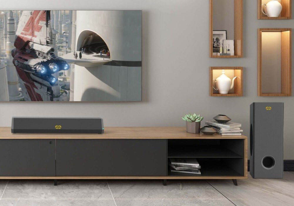 Na drewnianej szafce znajduje się soundbar z logo, obok stoi subwoofer z tym samym logo. Na ścianie wisi duży ekran z obrazem futurystycznego miasta i statku kosmicznego.