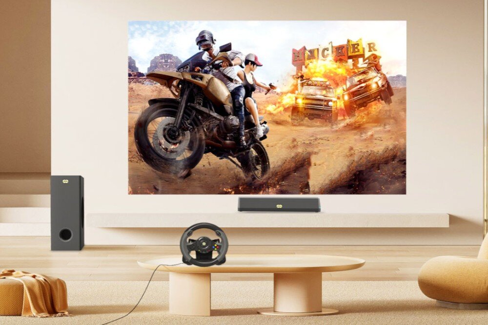 W salonie na ścianie wyświetlany jest dynamiczny obraz z motocyklistami i płonącym samochodem. Przed ekranem znajduje się soundbar i kierownica do gier, a obok subwoofer.