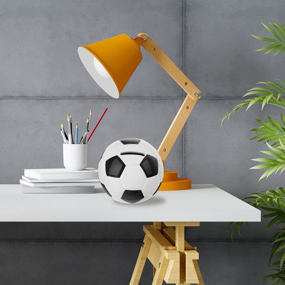 Na biurku znajduje się ceramiczna skarbonka w kształcie piłki nożnej oraz pomarańczowa lampa biurkowa. Obok skarbonki leży kubek z przyborami do pisania i kilka książek.