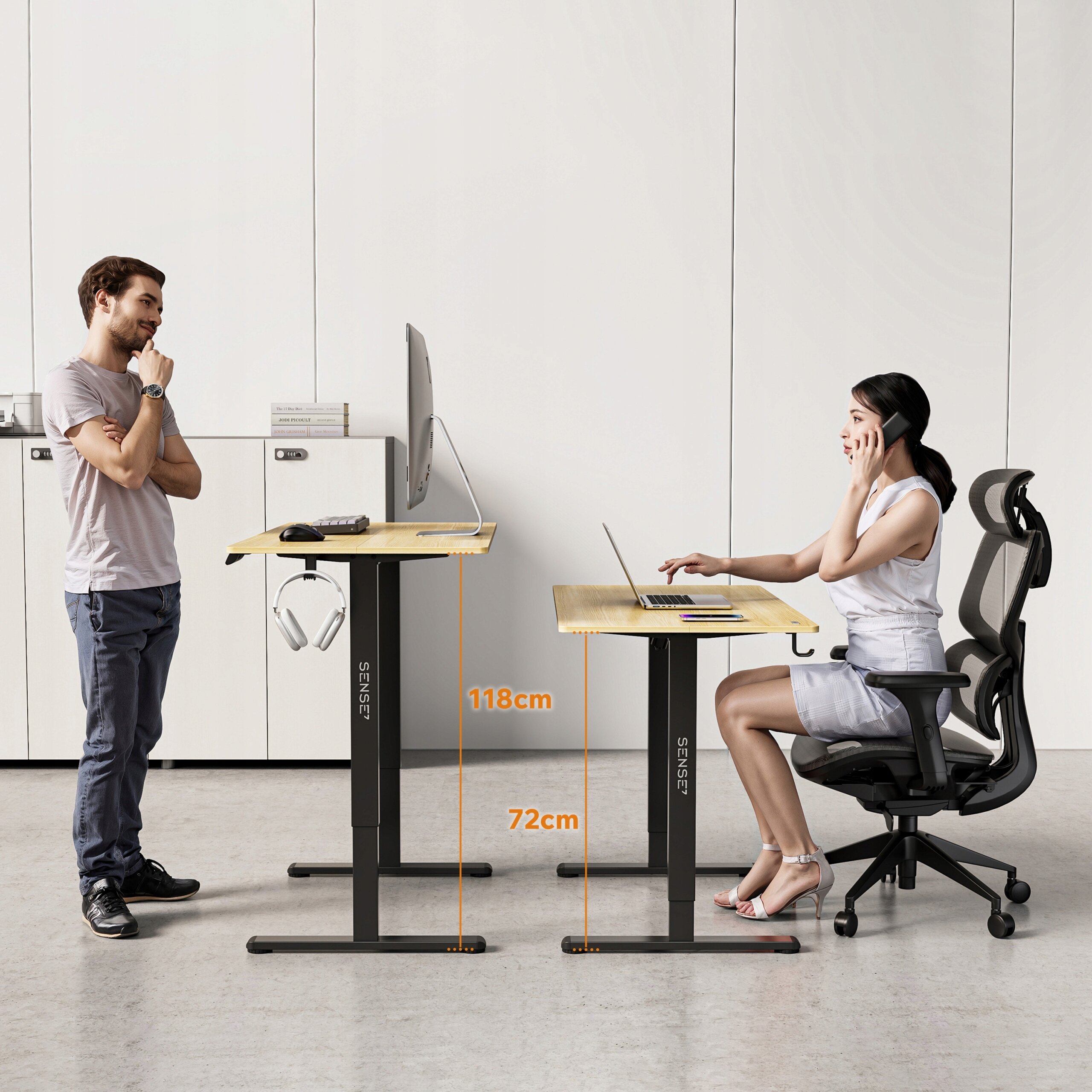 Na obrazie widoczne są dwa biurka z regulacją wysokości, jedno ustawione na 118 cm, a drugie na 72 cm, z logo SENSE7. Przy biurkach znajdują się osoby, jedna stoi, a druga siedzi na krześle biurowym.
