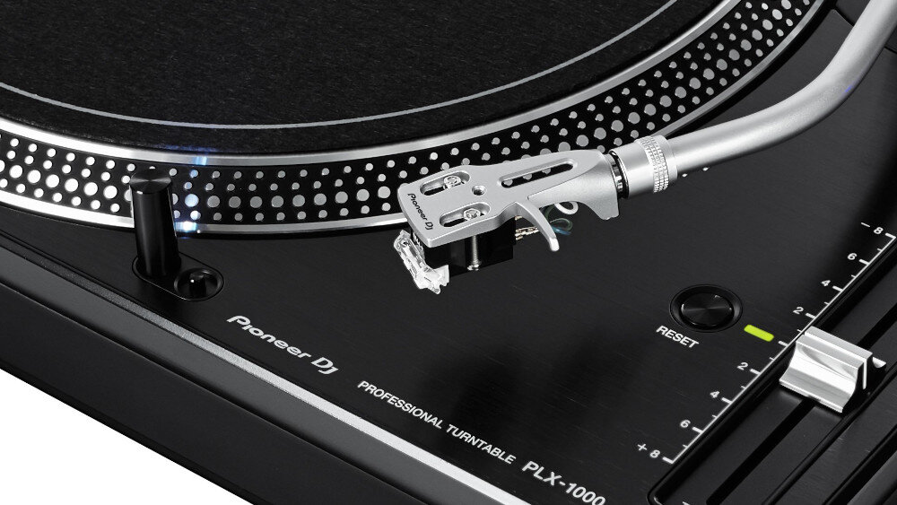 Zdjęcie przedstawia fragment gramofonu z widocznym ramieniem i igłą. Na obudowie znajduje się napis \'Pioneer DJ PROFESSIONAL TURNTABLE PLX-1000\'.