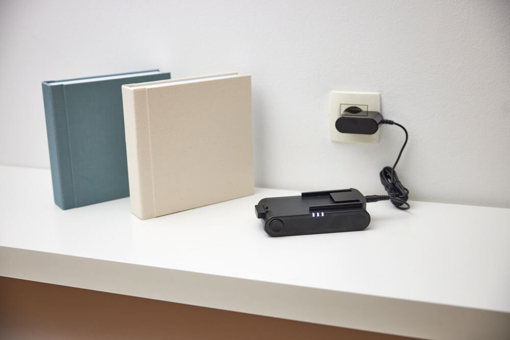 Na białej półce znajdują się dwa zamknięte albumy w kolorach niebieskim i beżowym oraz czarne urządzenie z podłączonym kablem do gniazdka elektrycznego. Urządzenie ma trzy świecące diody na obudowie.