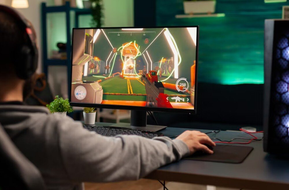 Osoba grająca w grę komputerową na monitorze, z widocznym kolorowym interfejsem gry. Obok monitora znajduje się klawiatura i myszka na podkładce.