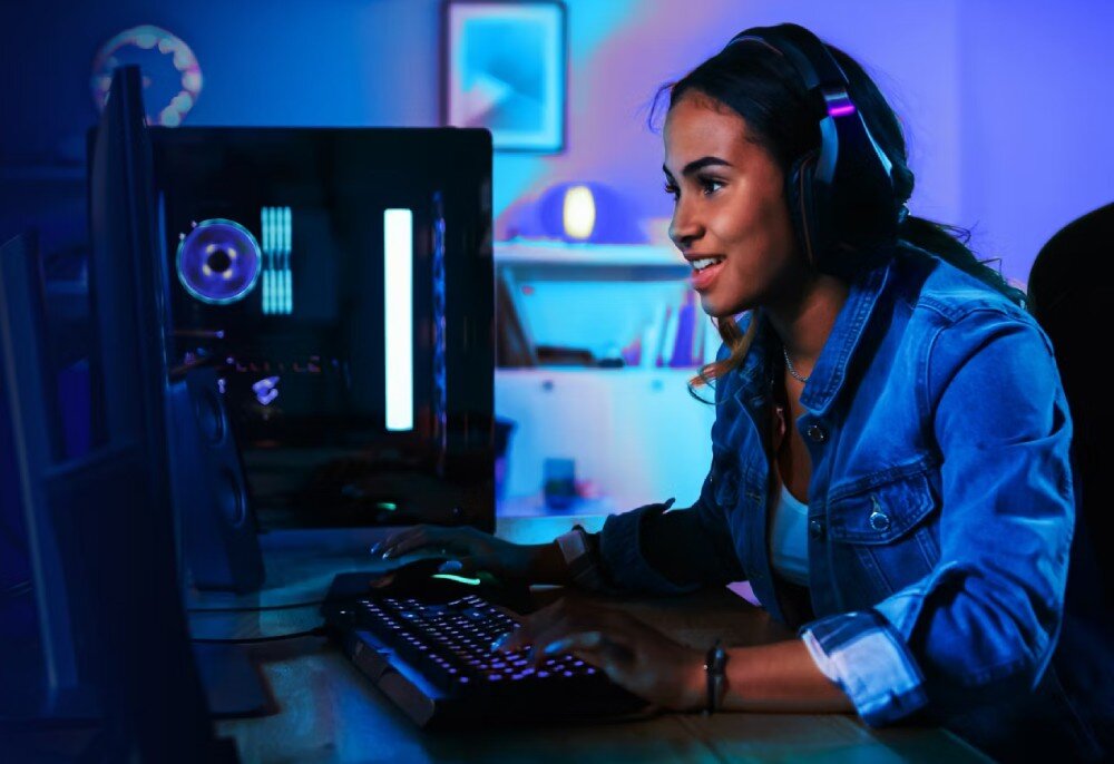 Osoba siedzi przy biurku, grając na komputerze z podświetlaną klawiaturą i słuchawkami na głowie. W tle widać półkę z książkami i lampką.