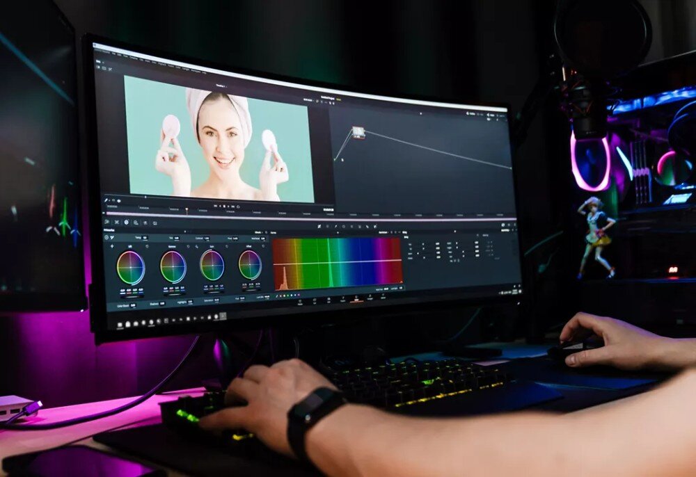 Osoba pracuje przy komputerze z dużym, zakrzywionym monitorem, na którym wyświetlany jest program do edycji wideo. Na ekranie widoczny jest obraz kobiety trzymającej dwa okrągłe przedmioty oraz narzędzia do korekcji kolorów.