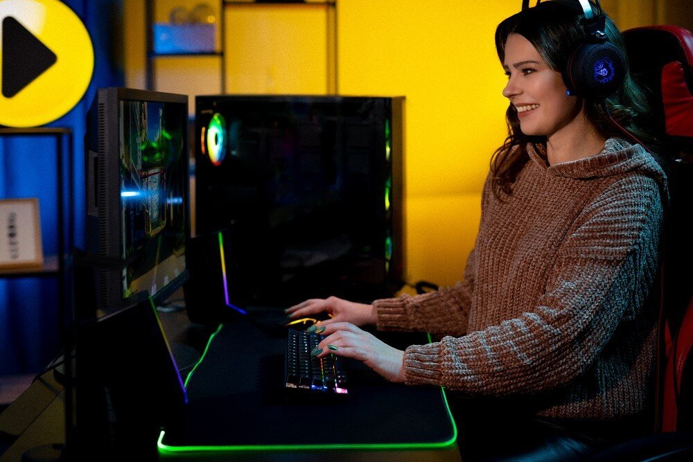 Komputer MAD DOG PHANTEKS-A02WR32WH Uśmiechnięta kobieta w brązowym swetrze i słuchawkach siedzi przy biurku przed monitorem komputera, trzymając dłonie na podświetlanej klawiaturze i myszce w ciemnym pokoju z kolorowym oświetleniem gamingowym