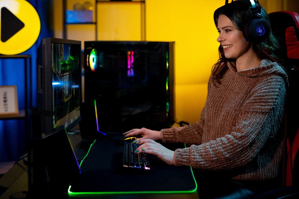 Komputer MAD DOG PHANTEKS-A04WR32WH Uśmiechnięta kobieta w brązowym swetrze i słuchawkach siedzi przy biurku przed monitorem komputera, trzymając dłonie na podświetlanej klawiaturze i myszce w ciemnym pokoju z kolorowym oświetleniem gamingowym