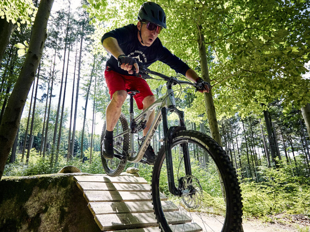 Rower górski MTB ROMET Dagger 2 29180130-B U18 29 cali Szaro-czarny las droga leśna osoba w kasku jedzie na rowerze zjeżdża z drewnianego mostka w tle niebo drzewa siodełko marki SELLE ITALIA typu Q-Bik wygodna pozycja podczas jazdy redukcja nacisku w kluczowych punktach sztyca DROPPER typu X-Fusion Manic Travel o długości 100 mm i średnicy mocowania 30,9 mm pozwala szybko zmienić wysokość siodełka