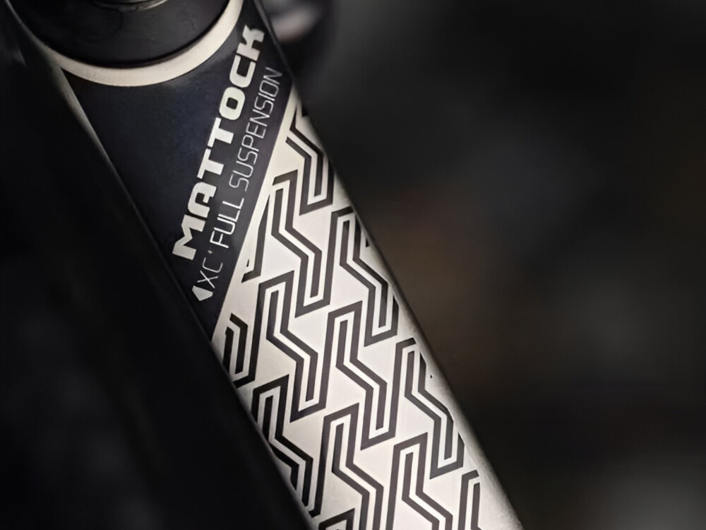 Zbliżenie na ramę roweru z widocznym wzorem geometrycznym i napisem \'MATTOCK XC FULL SUSPENSION\'.