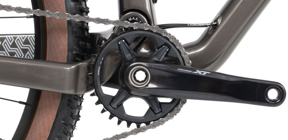 Zbliżenie na mechanizm korbowy roweru z widocznym napisem \'DEORE XT\'. Łańcuch jest zamontowany na zębatce, a rama roweru ma grafitowy kolor.