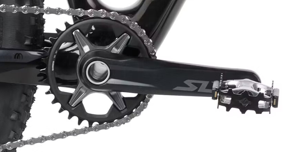 Zdjęcie przedstawia zbliżenie na mechanizm korbowy roweru z widocznym łańcuchem i pedałem. Na ramieniu korby znajduje się napis \'SLX\'.
