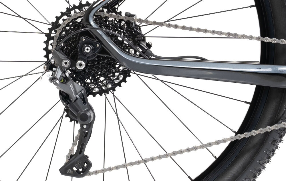 Zdjęcie przedstawia tylną część roweru z widocznym mechanizmem przerzutki i zębatkami. Rower ma czarny kolor i widoczny jest fragment opony oraz łańcucha.