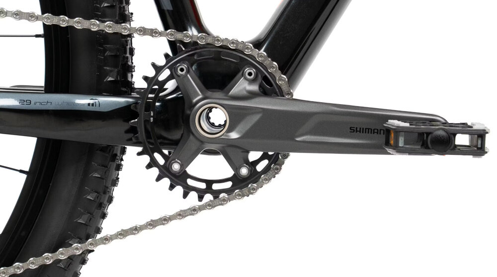 Zbliżenie na mechanizm korbowy roweru z widocznym napisem \'SHIMANO\' na ramieniu korby. Łańcuch jest napięty, a opona ma wyraźny bieżnik.