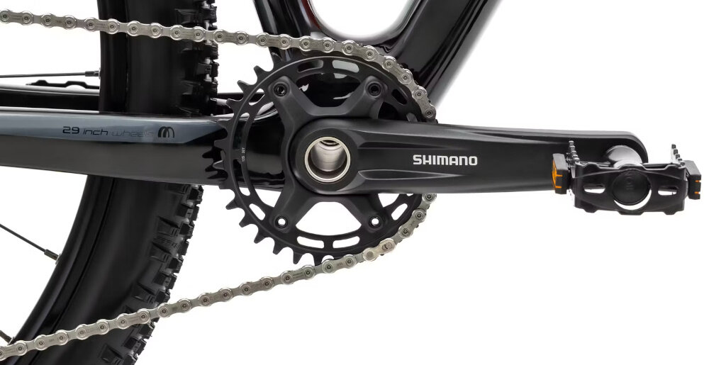 Zbliżenie na mechanizm korbowy roweru z widocznym napisem \'Shimano\' na ramieniu korby. Na ramie roweru widoczny jest napis \'29 inch\'.