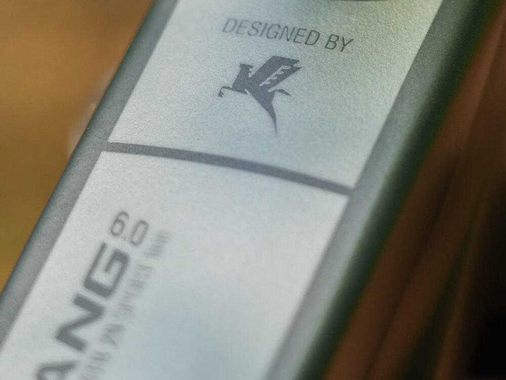 Zbliżenie na ramę roweru z widocznym napisem \'DESIGNED BY\' oraz logo w kształcie stylizowanego ptaka. Obok znajduje się napis \'ANG 6.0\'.