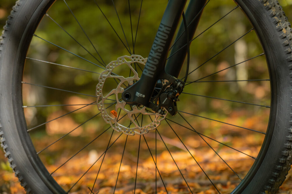Zbliżenie na przednie koło roweru górskiego z widocznym amortyzatorem z napisem \'ROCKSHOX\' i tarczą hamulcową. W tle rozmyte liście na ziemi w jesiennych kolorach.