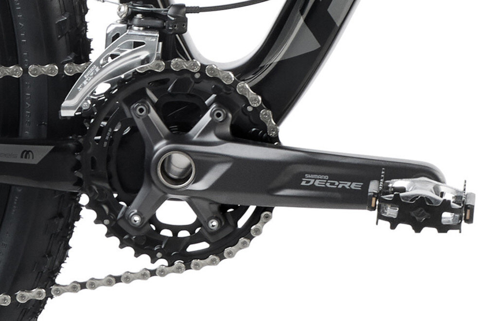 Zdjęcie przedstawia mechanizm korbowy roweru z widocznym napisem \'Shimano Deore\'. Łańcuch jest nałożony na zębatkę, a pedał jest przymocowany do ramienia korby.