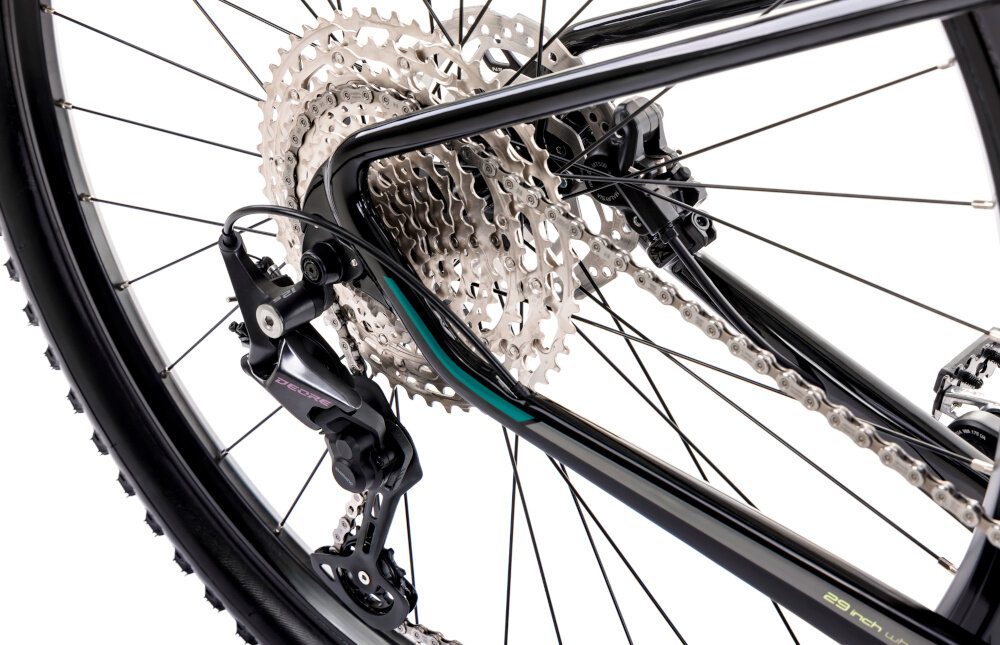 Rower górski MTB ROMET Mustang EVO 3 29170148-A U17 29 cali Czarno-zielony zbliżenie na tylną przerzutkę widok pod skosem białe tło przerzutki marki SHIMANO 12 przełożeń duża wytrzymałość i sprawne działanie zmiana biegów jest płynna i bezproblemowa tylne przerzutki SHIMANO Deore typu RD-M6100 trwałość stabilność i precyzja precyzyjna praca mechanizmu