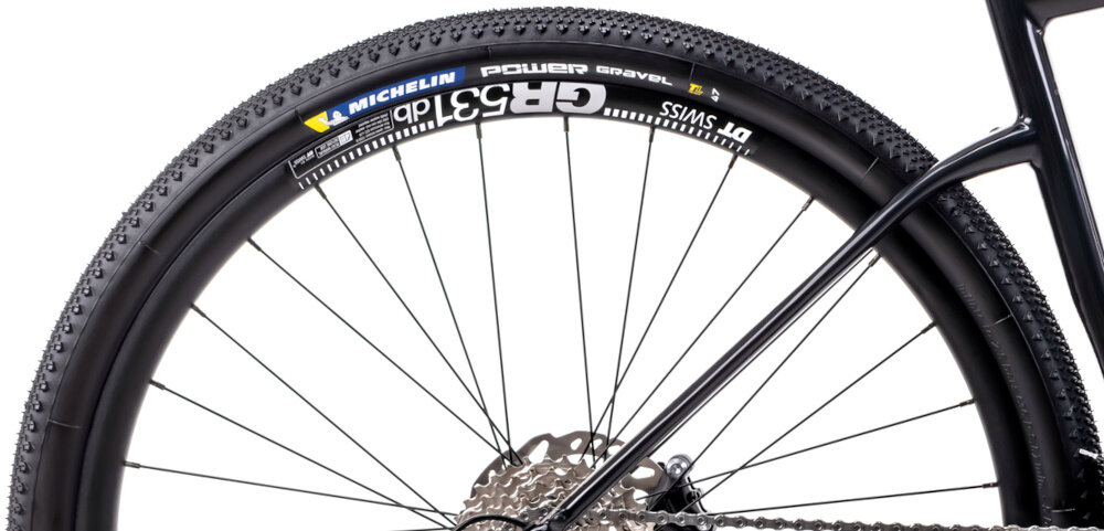 Rower gravel ROMET NYK 2.0 28460099-A U17 28 cali Fioletowy zbliżenie na tylne koło białe tło opony marki MICHELIN w rozmiarze 28 cali głęboki bieżnik skutecznie wspiera jazdę stabilny tor jazdy aluminiowe obręcze marki DT Swiss GR 531 sztywność niska masa piasty SHIMANO HB-RS470 w standardzie 12x100 mm z przodu i 12x142 mm z tyłu sztywność kół regularne sprawdzanie ciśnienia powietrza wartość na bocznej części opony