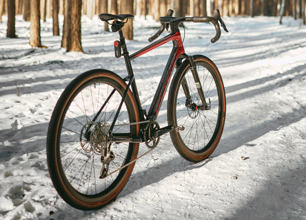 Rower gravel ROMET NYK 3.0 SRAM 28540363-A U21 28 cali męski Czerwono-czarny zimowa sceneria rower widoczny od przodu pod skosem ustawiony na śniegu w lesie stalowy łańcuch marki ROMET konstrukcja dostosowana do różnych napędów  przenoszenie mocy wysoka odporność na zużycie