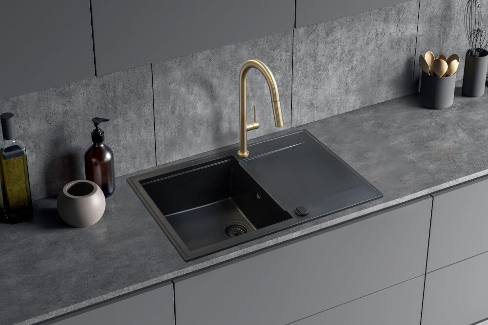 Na obrazie widoczny jest nowoczesny zlew kuchenny z czarną komorą i złotą baterią. Na blacie znajdują się różne akcesoria kuchenne, w tym dozownik na mydło i pojemnik na sztućce.