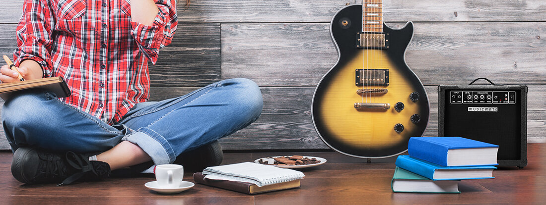 Osoba siedzi na podłodze z notesem, obok znajduje się gitara elektryczna i wzmacniacz z logo \'musicmate\'. Na podłodze leżą książki, filiżanka i talerz z ciastkami.