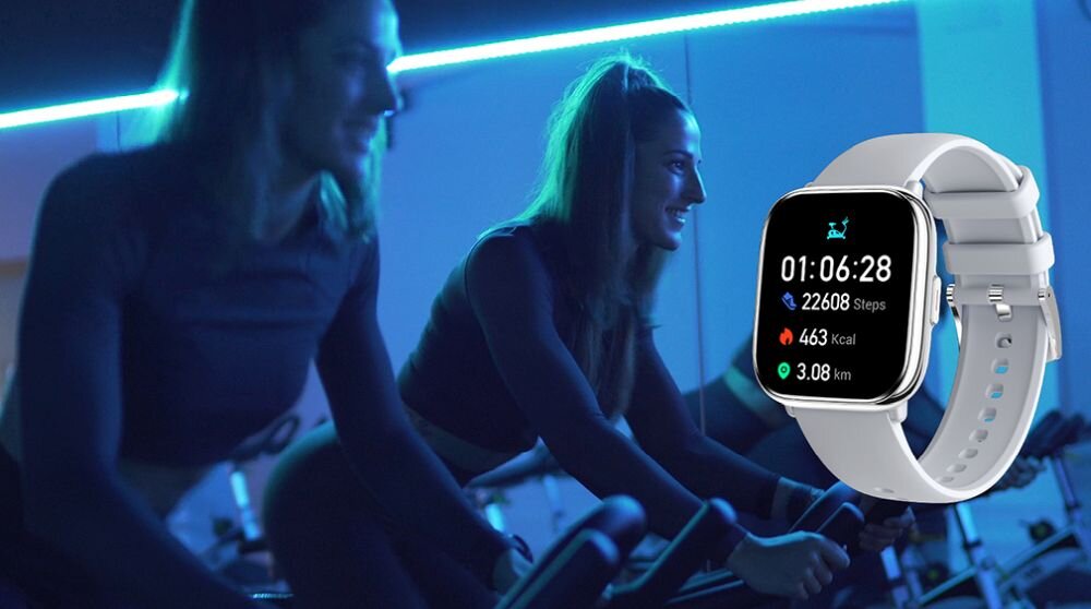 Smartwatch z wyświetlaczem pokazującym czas 01:06:28, 22088 Steps, 463 Kcal, 3.08 km. W tle osoby na rowerach treningowych w oświetleniu neonowym.