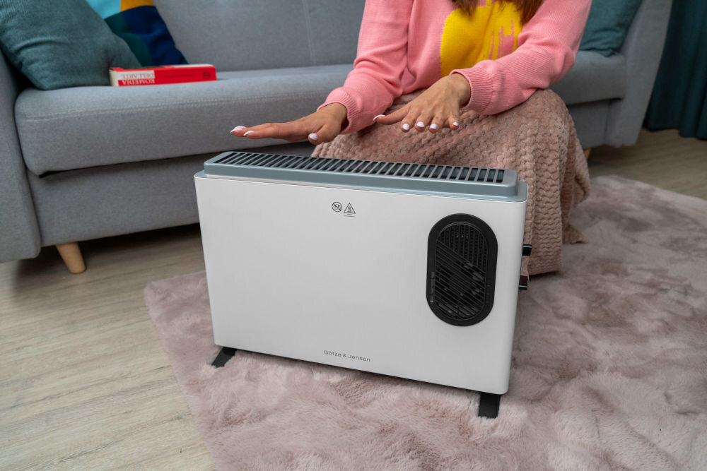 Na dywanie stoi biały grzejnik konwektorowy z czarnym panelem bocznym i widocznym napisem \'GÖTZE & JENSEN\'. W tle znajduje się szara sofa, a na niej leży czerwona książka.