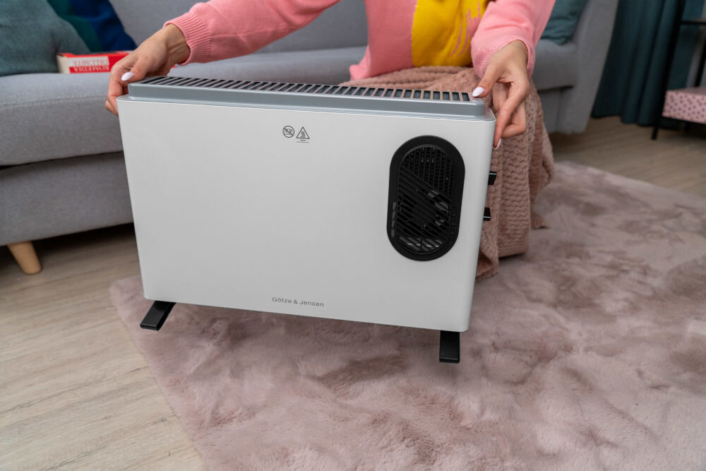 Osoba trzyma biały grzejnik konwektorowy z czarnym wentylatorem z boku. Na grzejniku widoczne jest logo \'götze & jensen\'.