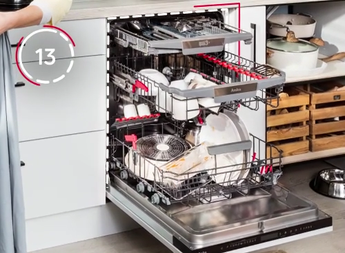 Otwarta zmywarka z widocznymi trzema poziomami koszy, wypełnionymi naczyniami i sztućcami. Na froncie zmywarki widoczny jest napis \'Amica\'.