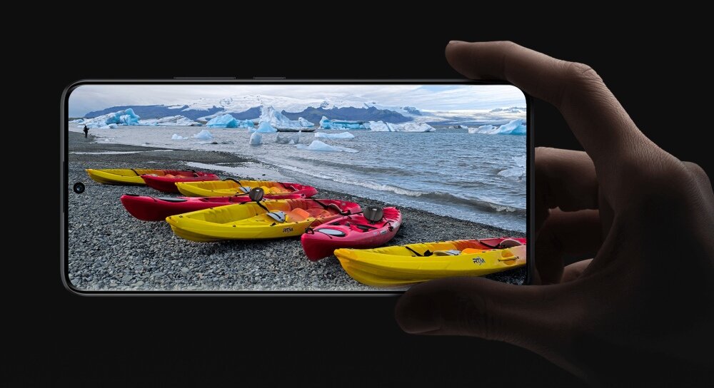 Osoba trzyma smartfon, na ekranie którego wyświetla się zdjęcie plaży z kilkoma kajakami w różnych kolorach, a w tle widać morze i góry lodowe. Tło obrazu jest czarne, co podkreśla wyświetlacz urządzenia.