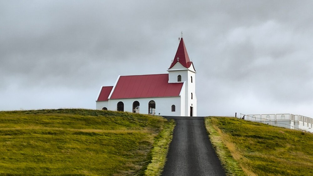 Na obrazie widoczny jest biały kościół z czerwonym dachem, stojący na wzgórzu. W tle niebo jest zachmurzone.