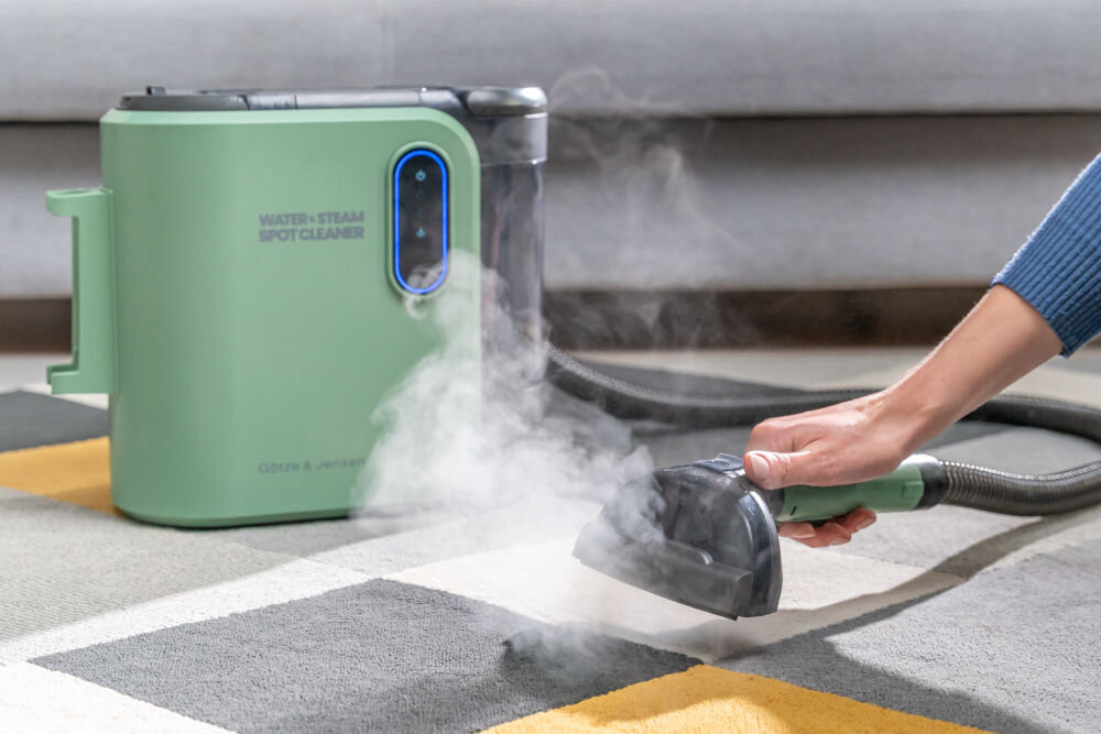Na obrazie widoczny jest zielony odkurzacz piorący z napisem \'WATER+STEAM SPOT CLEANER\' oraz \'GÖTZE & JENSEN\'. Obok urządzenia znajduje się ręka trzymająca dyszę, z której wydobywa się para, skierowana na dywan.