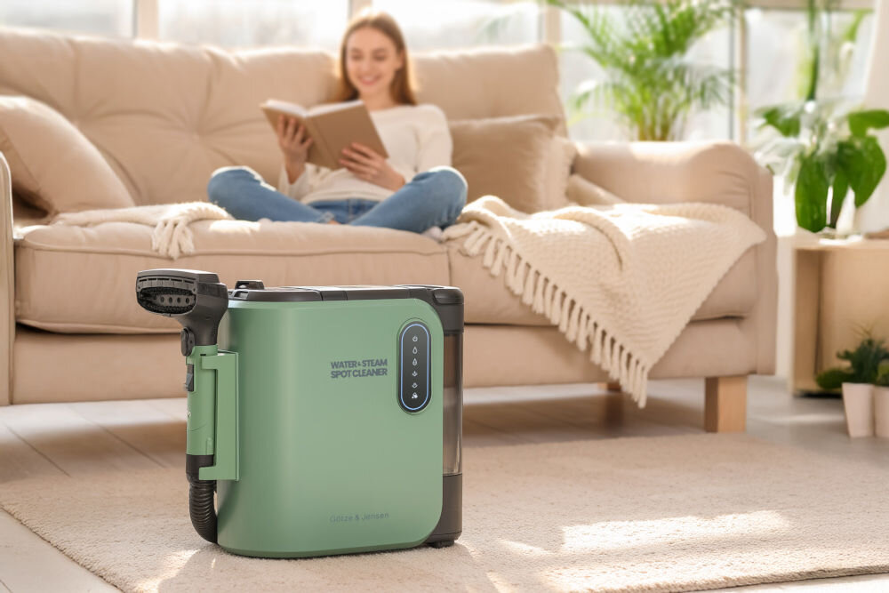 Na pierwszym planie znajduje się odkurzacz piorący w kolorze zielonym z napisem \'WATER-STEAM SPOT CLEANER\'. W tle widać osobę siedzącą na sofie z książką w rękach.