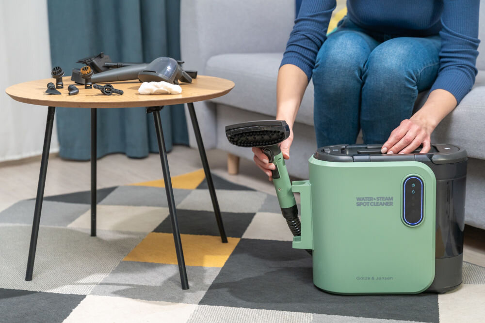 Osoba trzyma zielony odkurzacz piorący z napisem \'WATER-STEAM SPOT CLEANER\' oraz \'Götze & Jensen\'. Na stoliku obok znajdują się różne akcesoria do odkurzacza.