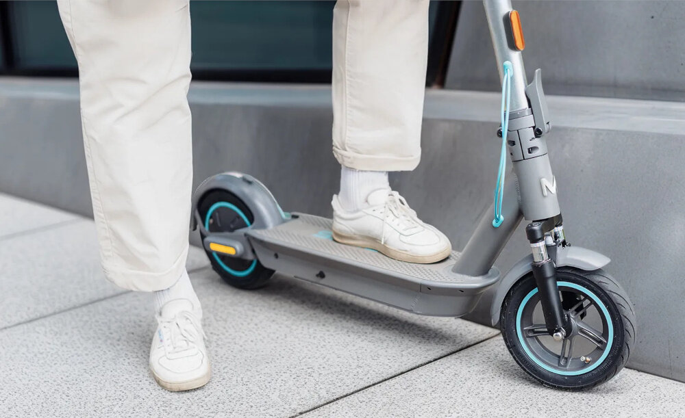 Hulajnoga elektryczna MOTUS Scooty 10 Plus Gen 6 65km 500W Amortyzacja 10 cali Szary chodnik zbliżenie na deck osoba opiera jedną stopę o deck wytrzymała konstrukcja z wysokiej jakości aluminium waga 20,5 kg dla osób o maksymalnej wadze do 120 kg spory deck szerokość 17 cm komfortowa i stabilna jazda antypoślizgowa powłoka