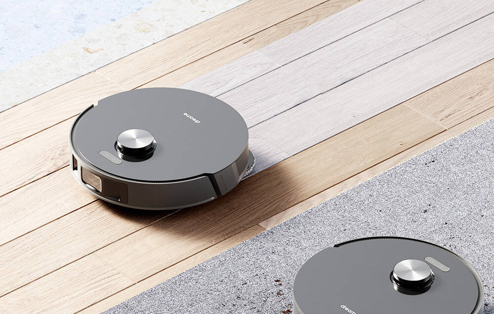 Na podłodze znajdują się dwa okrągłe roboty sprzątające w kolorze szarym. Na jednym z nich widoczny jest napis \'dreame\'.